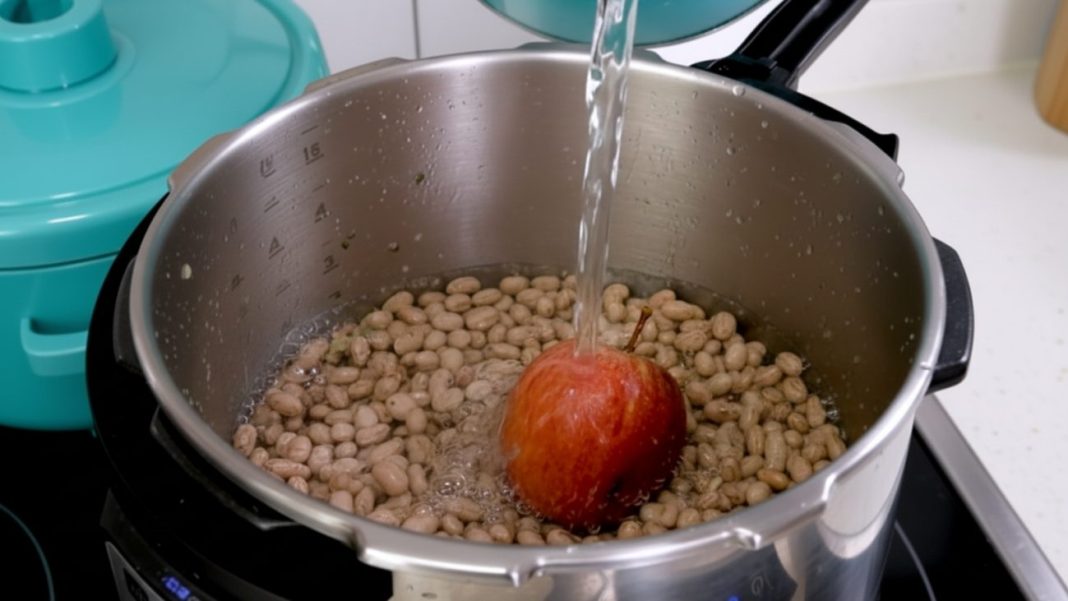 Panela de pressão com feijão e uma maçã inteira sendo cobertos por água, ilustrando a técnica de cozinhar feijão com uma maçã junto para reduzir gases e amaciar os grãos